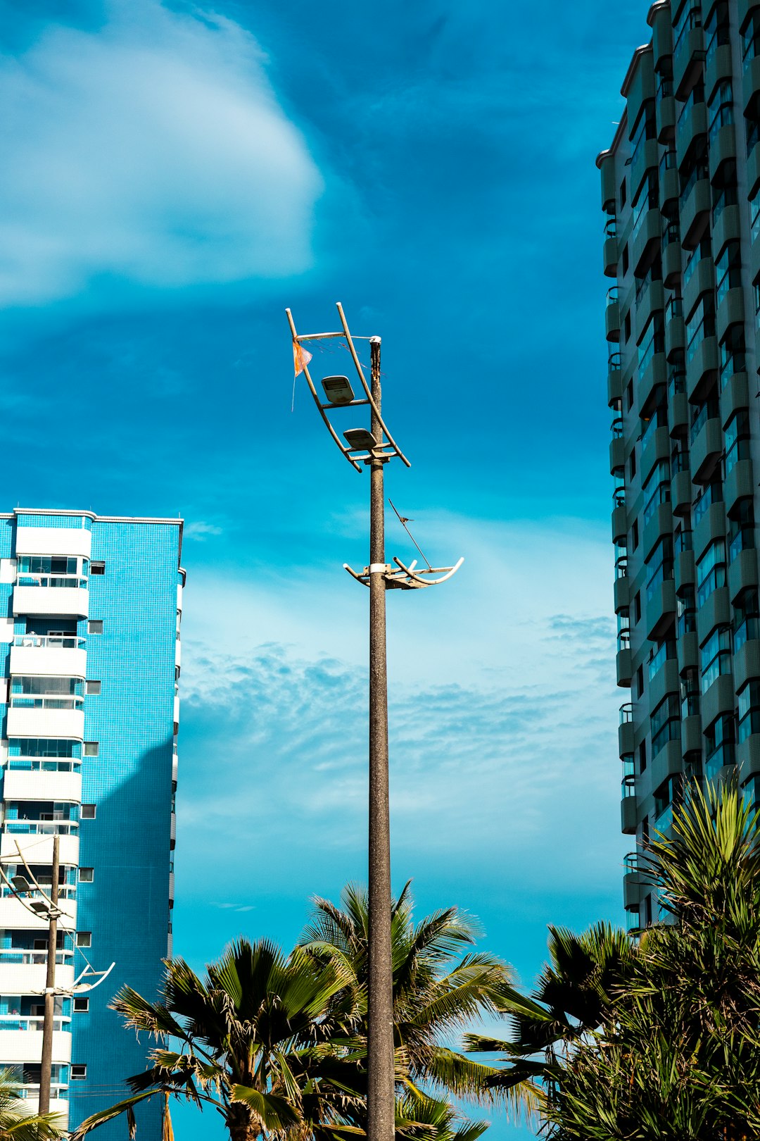 services-03 A tall, weathered lamppost stands between two modern apartment buildings under a bright blue sky with scattered clouds. Palm trees frame the bottom of the image, adding a tropical feel to the urban landscape.
