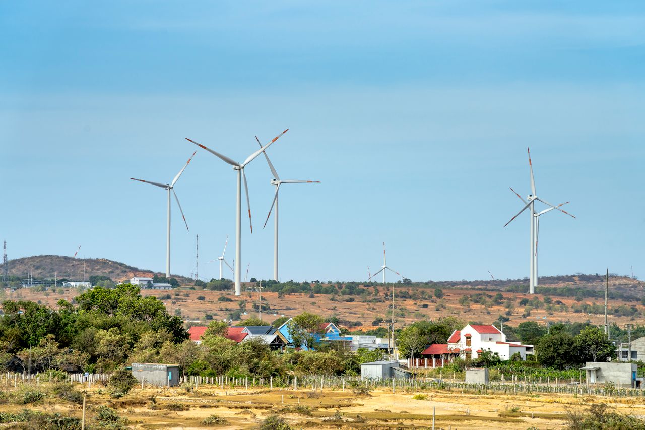 gallery-04 Scenic view of wind generators on land between buildings and mount under cloudy blue sky